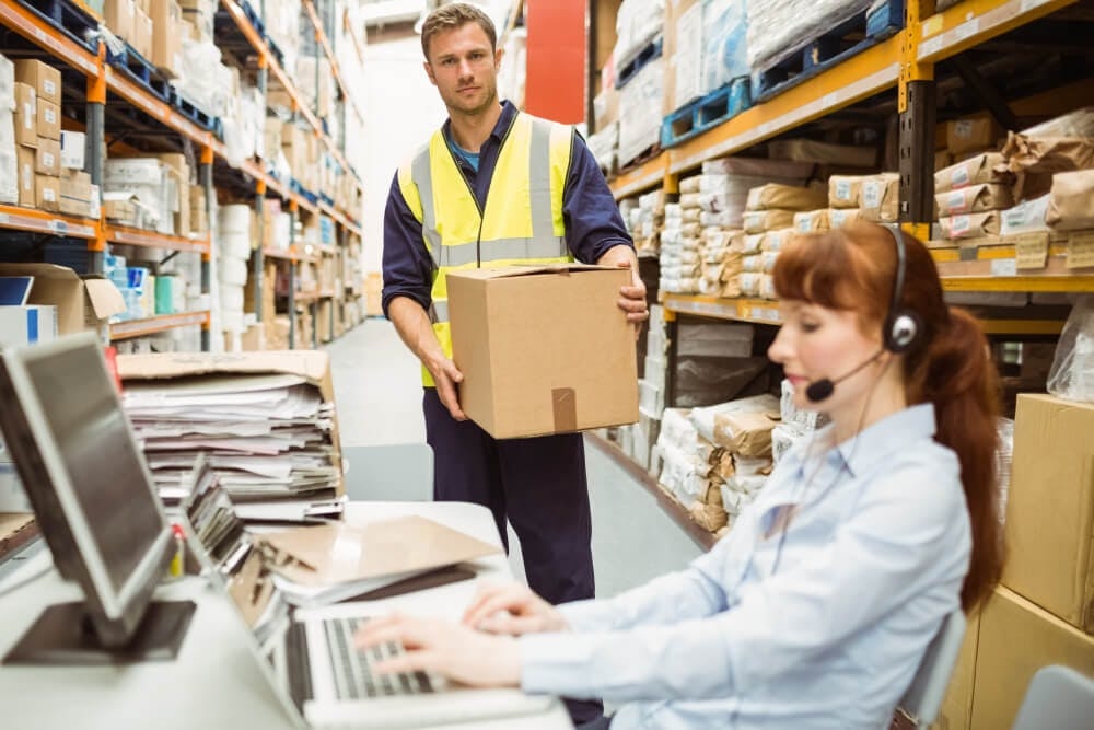Usługa logistyczna – magazynowanie towarów. Kobieta pracująca przy komputerze w magazynie wysokiego składowania oraz mężczyzna trzymający karton